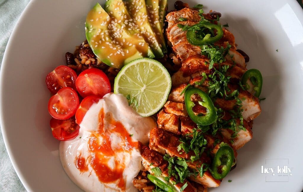 Chicken burrito bowl topped with sliced chicken, avocado, beans, sweetcorn, cheddar, and chilli cream drizzle.