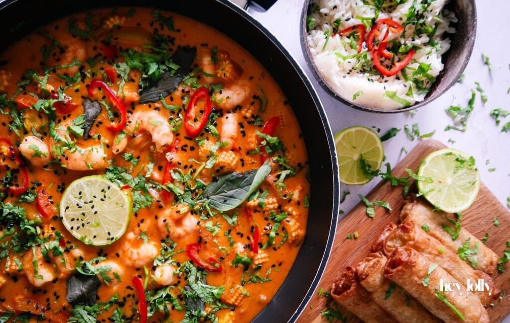 Overhead shot of fresh ingredients for Thai Red King Prawn curry including prawns, baby corn, red pepper, and courgette on a wooden board.