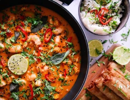 Overhead shot of fresh ingredients for Thai Red King Prawn curry including prawns, baby corn, red pepper, and courgette on a wooden board.