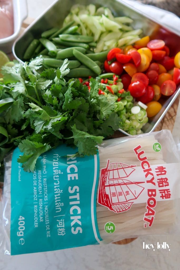Fresh ingredients for Thai poached chicken salad laid out: glass noodles, cherry tomatoes, cucumber, sugar snap peas, herbs, lime, and coconut milk.