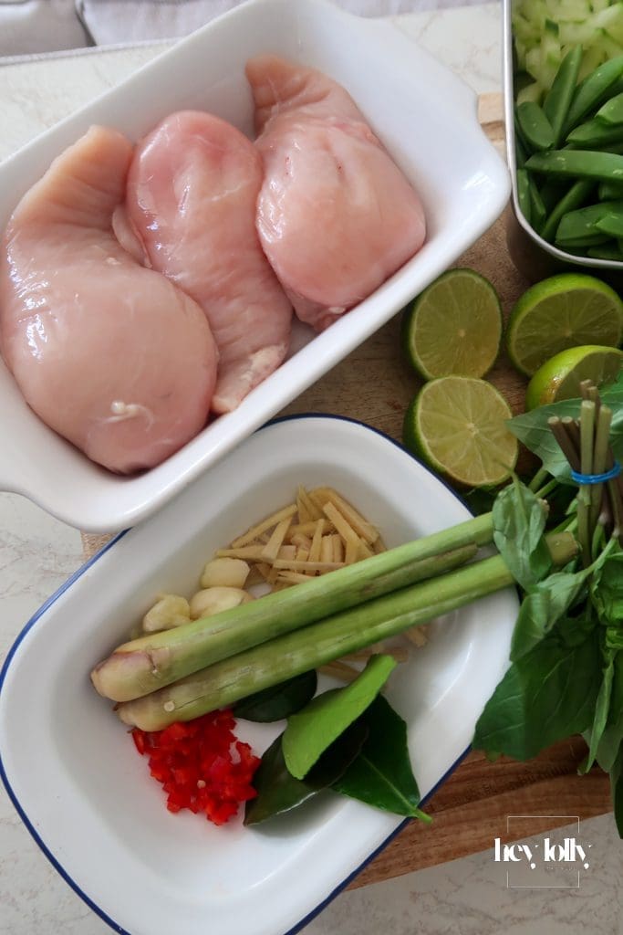 Fresh ingredients for Thai poached chicken salad laid out: glass noodles, cherry tomatoes, cucumber, sugar snap peas, herbs, lime, and coconut milk.