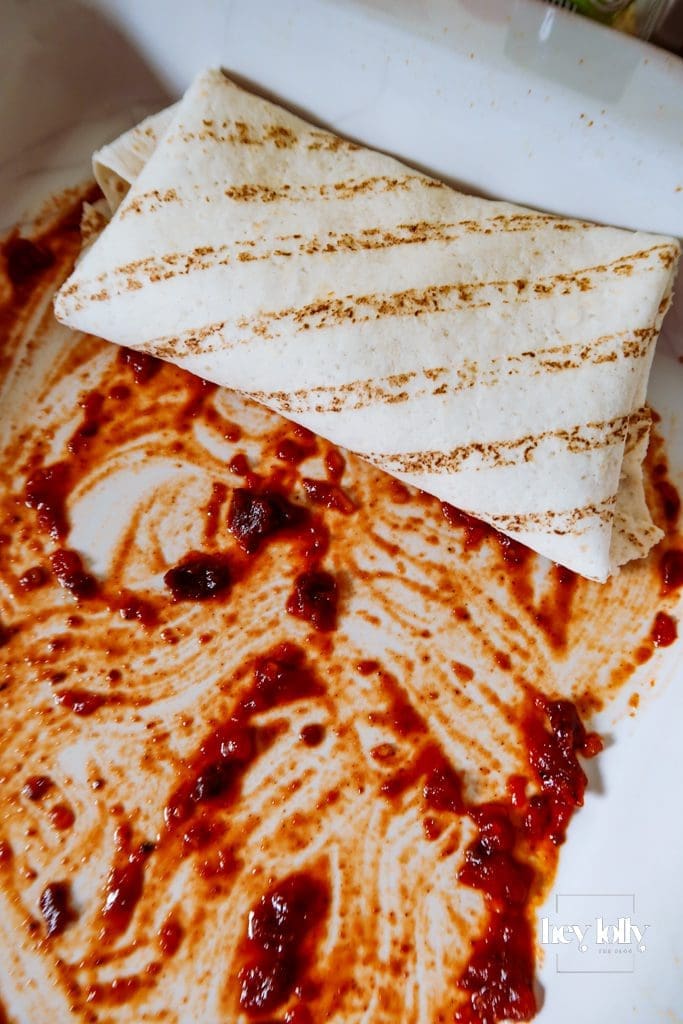 Rolling tortillas filled with corn and butter bean mixture on the worktop, sauce splatters visible around.