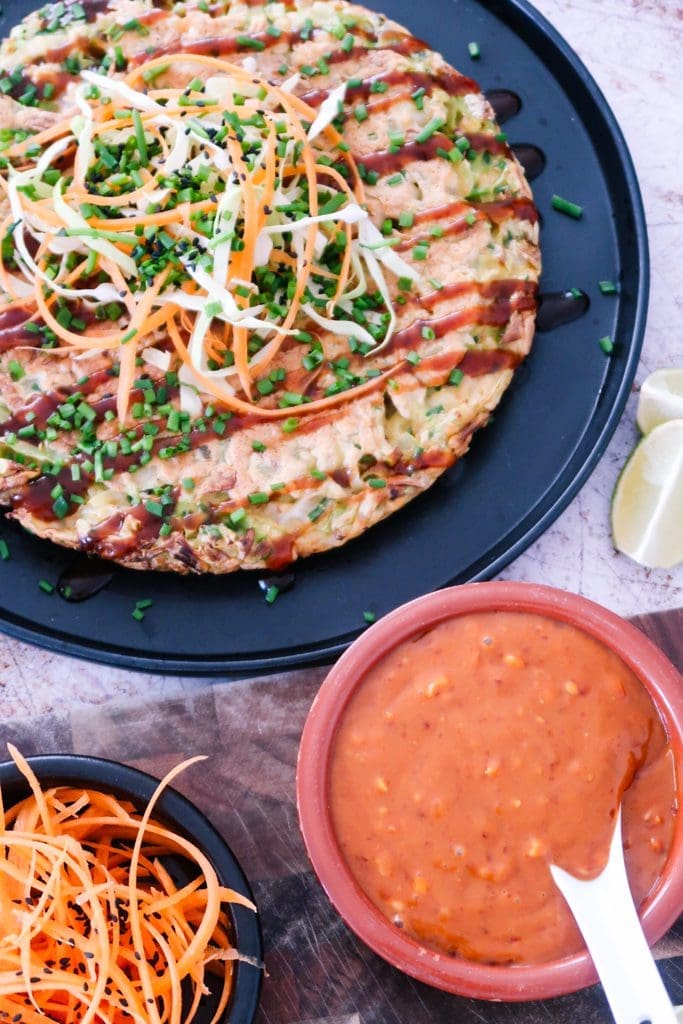 overhead shot of okonomiyaki pancake with gochujang, spring onions and sesame seeds on a white rustic table