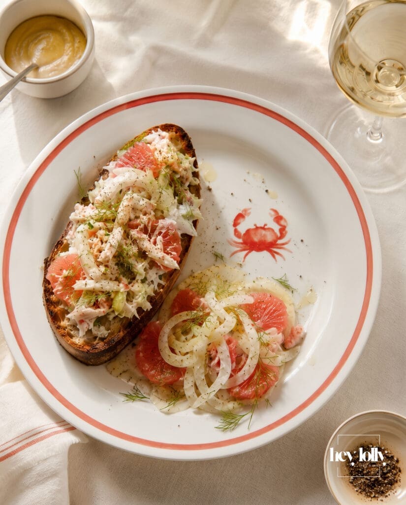 Crab toast with fennel and grapefruit salad on coral-rimmed crab plates with white wine and ivory linen table styling.