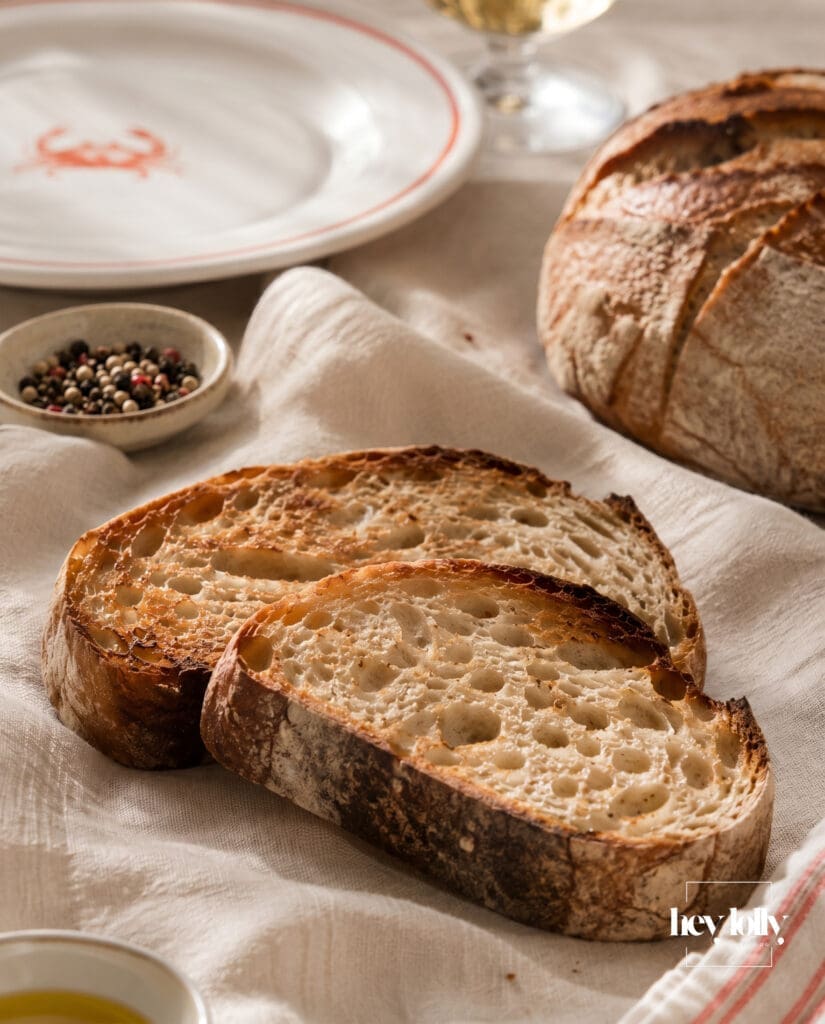 Golden toasted artisan sourdough slices on ivory linen beside coral-rimmed crab plate.