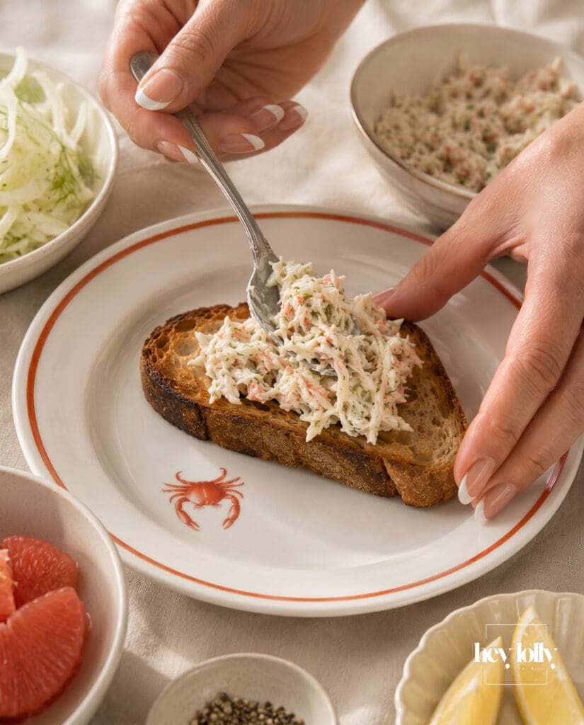Female hands with French tip manicure assembling crab toast with fennel and grapefruit salad on toasted sourdough.