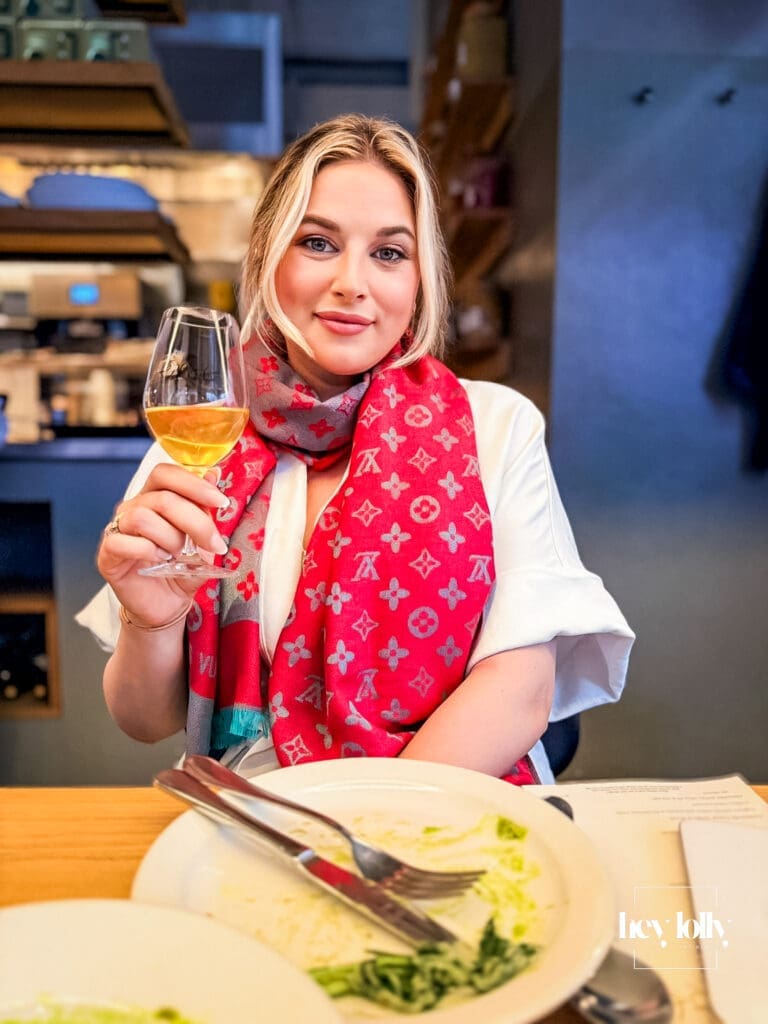 Blonde woman seated at the bar inside Erst Manchester holding a cocktail, wearing a white blouse and colourful scarf in a softly lit modern restaurant interior – Erst Manchester review