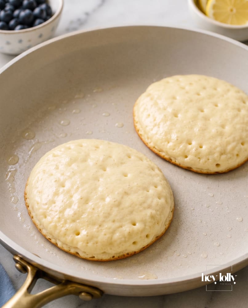 Lemon ricotta pancakes cooking in a frying pan with small bubbles forming on the surface before flipping.