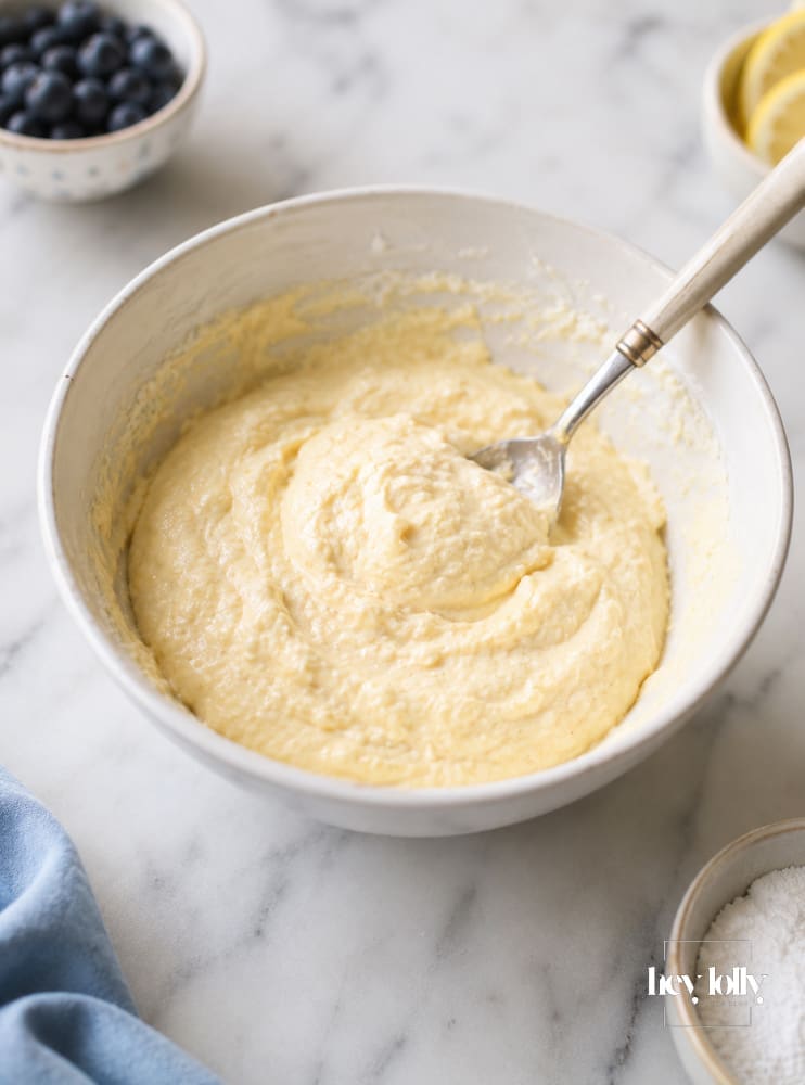Thick lemon ricotta pancake batter in a white mixing bowl with blueberries, lemon slices and sugar on a pale marble surface.