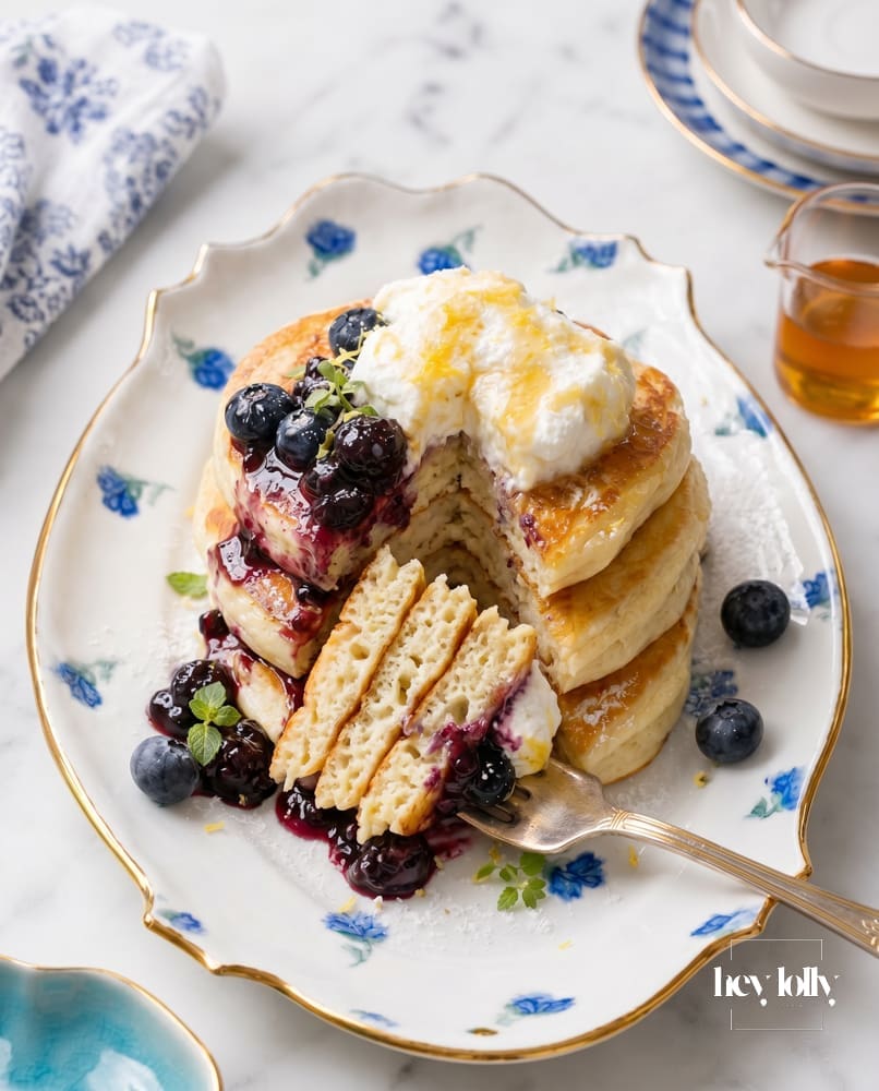 Close-up of fluffy lemon ricotta pancakes with glossy blueberry compote, soft cream, icing sugar and fresh lemon zest.