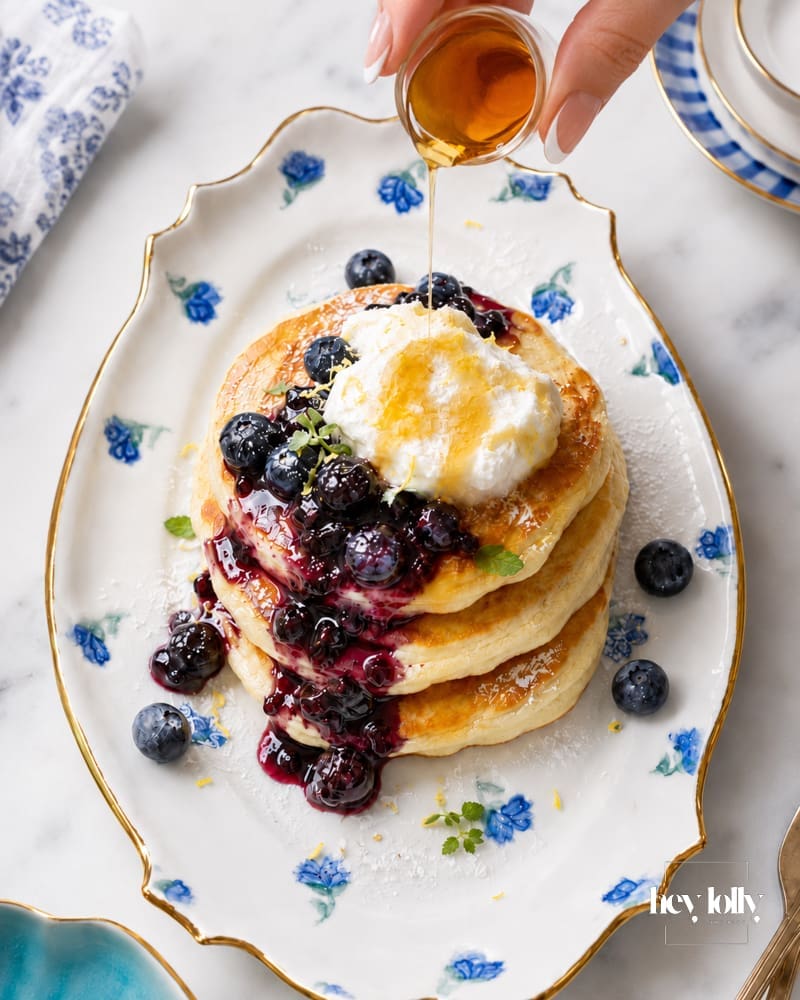 Female hand with almond French manicure pouring syrup over lemon ricotta pancakes with blueberry compote and cream.